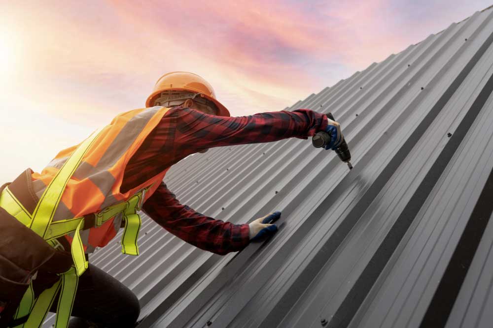 Korrect Roofing contractor in safety gear using a drill to install durable metal roofing panels during sunset.