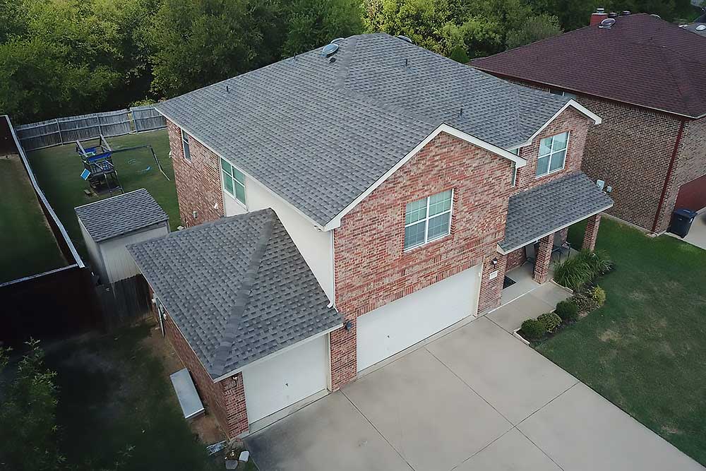 Aerial view of a two-story brick house with a gray shingle roof and a double garage. The property is surrounded by a green lawn and trees, ideal for Fort Worth roofing services, showcasing a well-maintained residential area.| Korrect General Contracting Fort Worth TX