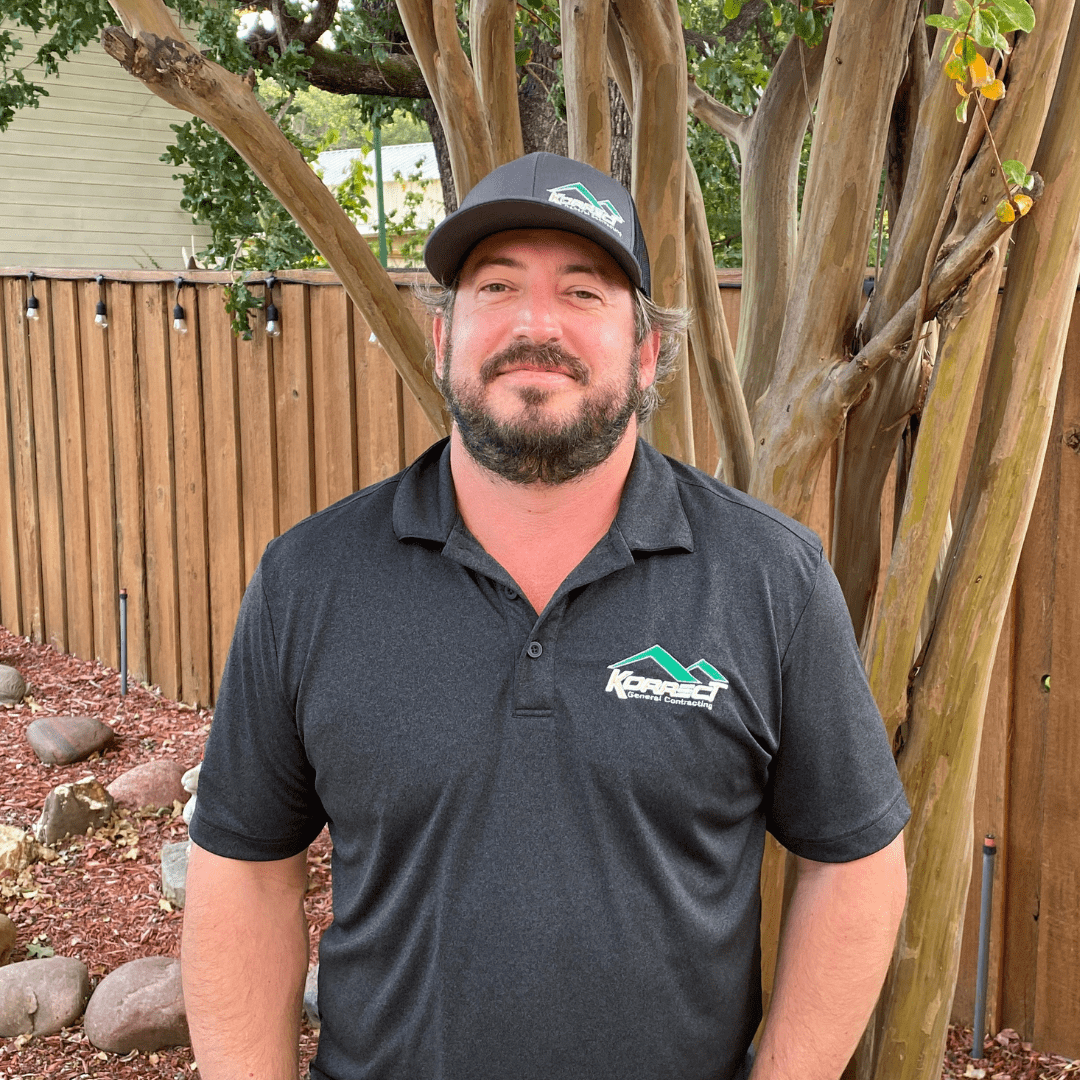 A man in a black polo shirt with a company logo stands in front of a wooden fence and trees. He appears confident, embodying the professionalism of a Fort Worth roofing contractor.| Korrect General Contracting Fort Worth TX