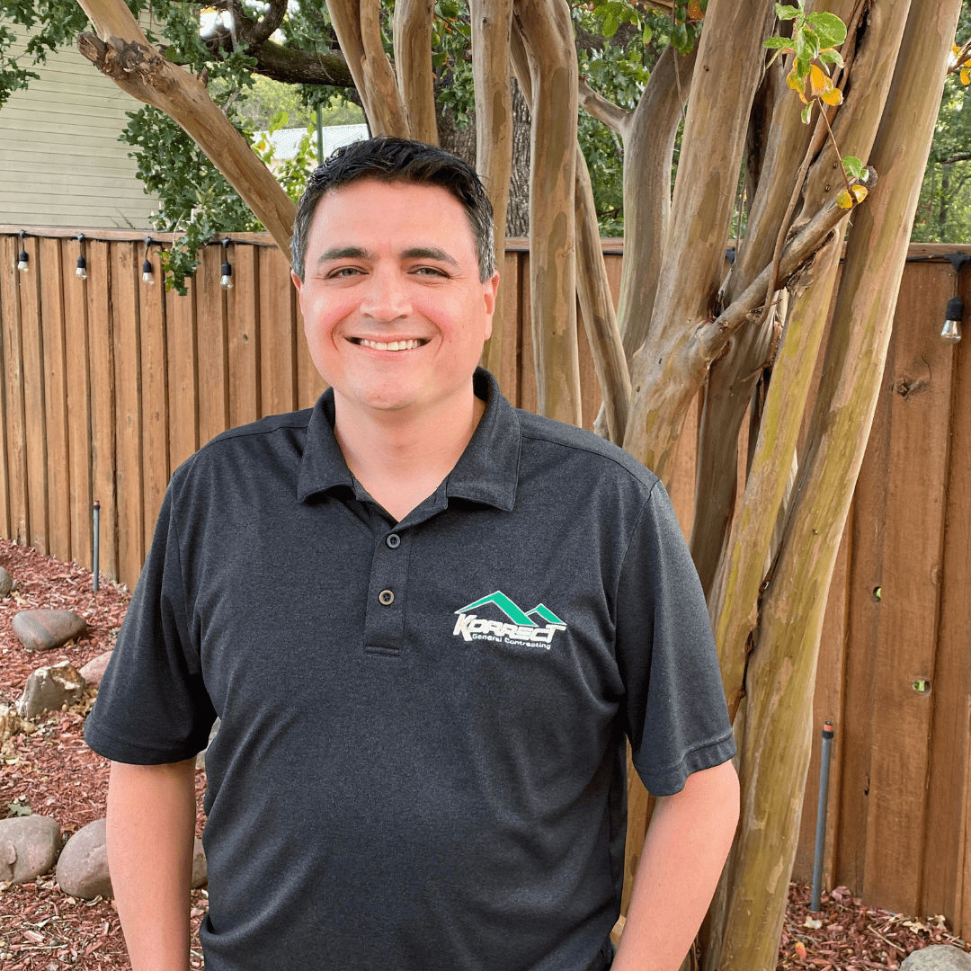 A smiling man in a dark polo shirt with a company logo stands in front of a wooden fence and tree. The setting sun casts a warm glow, highlighting the garden. This image represents a professional from a Fort Worth roofing contractor.| Korrect General Contracting Fort Worth TX