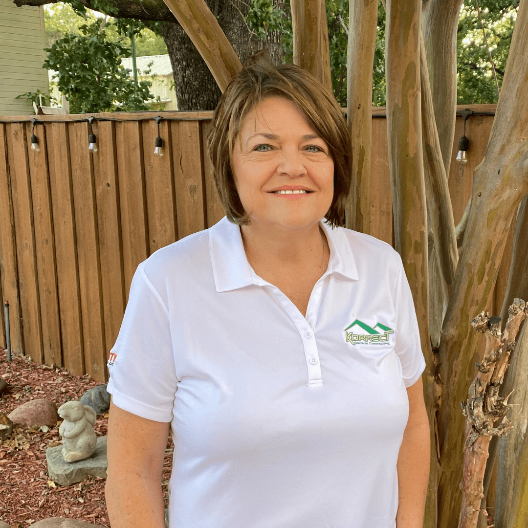A woman in a white polo stands smiling in a garden with a wooden fence and trees. Her shirt features a Korrect General Contracting logo, highlighting services like Fort Worth roofing and general contracting in the Dallas Fort Worth area.| Korrect General Contracting Fort Worth TX