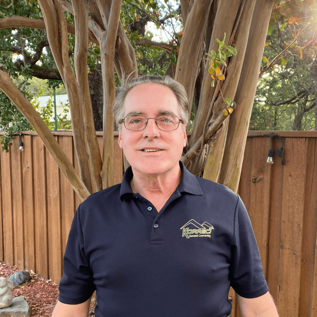 A man in a navy Korrect General Contracting shirt stands in front of a tree and wooden fence. He smiles at the camera, surrounded by greenery. The scene reflects a professional Fort Worth roofing environment.| Korrect General Contracting Fort Worth TX