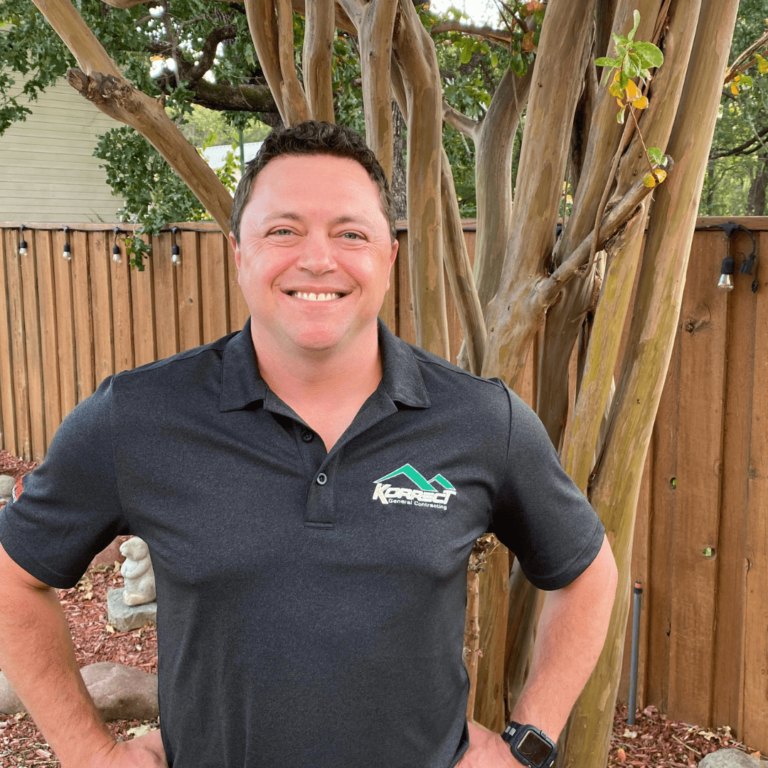 A smiling man in a black polo with a company logo stands in a garden with a wooden fence and trees in the background, representing a Fort Worth roofing contractor specializing in roof repair and inspections.| Korrect General Contracting Fort Worth TX