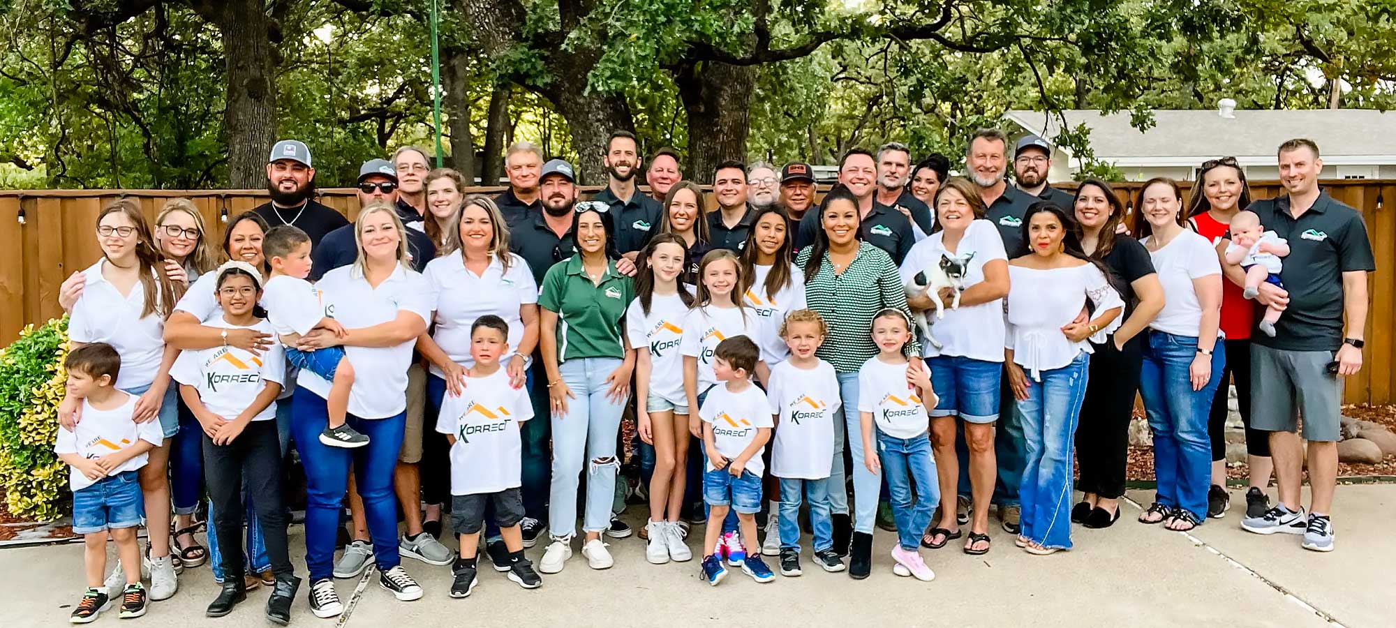 A large group of people of various ages, including children and adults, stand together outdoors, smiling in a casual setting with trees and a wooden fence behind them. This gathering represents a Fort Worth roofing community event.| Korrect General Contracting Fort Worth TX