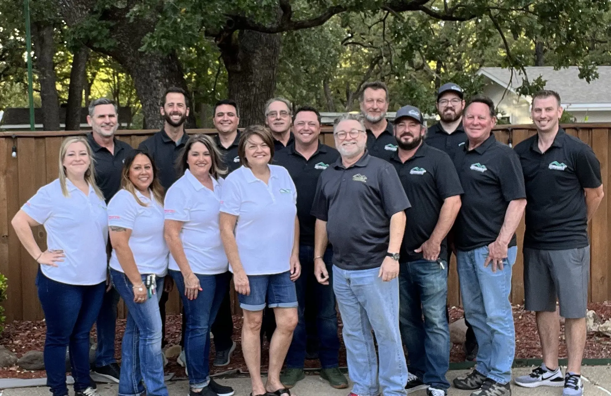 A group of men and women wearing matching company shirts stand outdoors in front of a wooden fence, smiling at the camera. The team represents Korrect General Contracting, a DFW contractor specializing in Fort Worth roofing and other services.| Korrect General Contracting Fort Worth TX