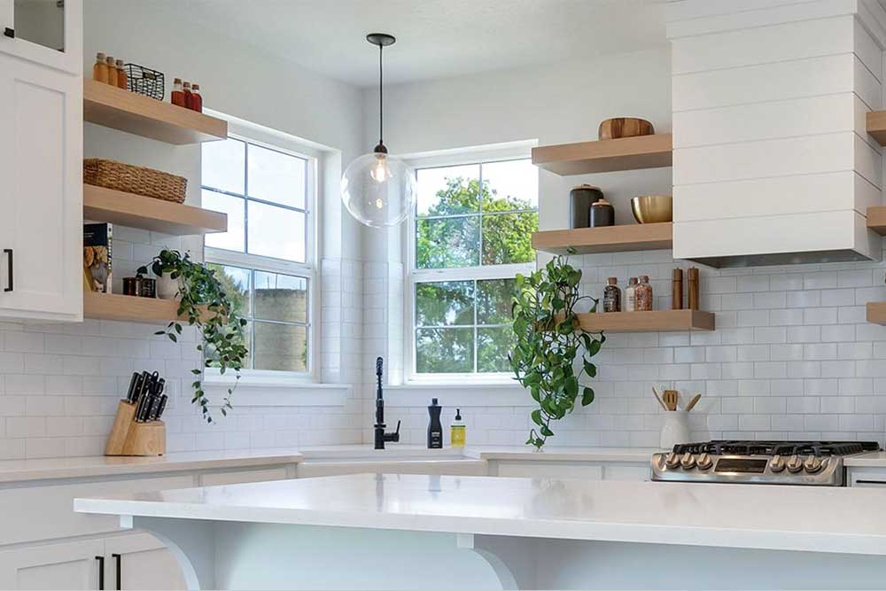 A modern kitchen with white cabinets, open wooden shelves, and green plants on a tiled backsplash. Large windows provide natural light, highlighting the spacious countertop, ideal for Dallas Fort Worth kitchen remodeling projects.| Korrect General Contracting Fort Worth TX